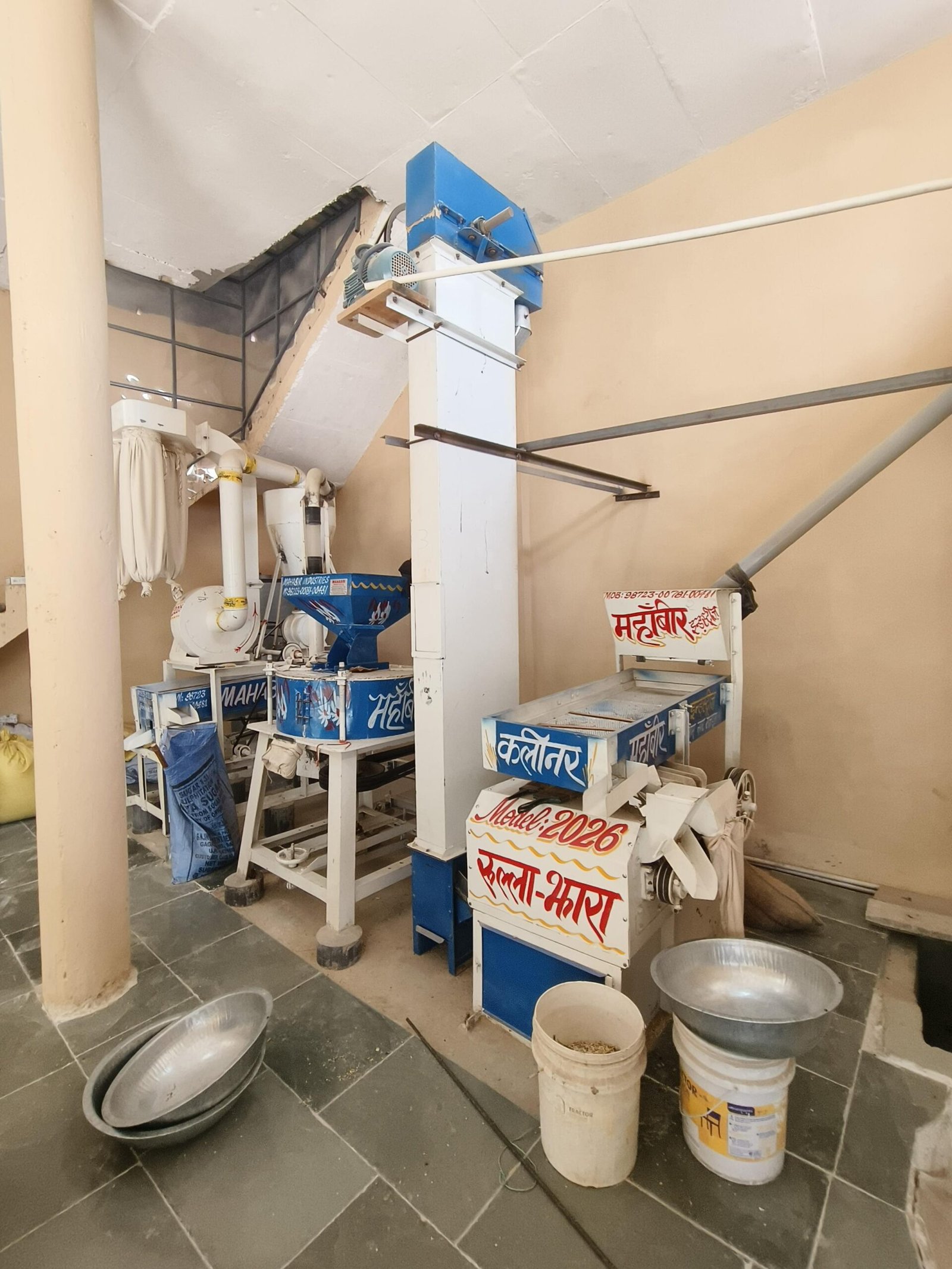 Industrial processing equipment in a workshop: blue control unit atop a white column, with belts, pipes, and metal basins/buckets on a tiled floor.