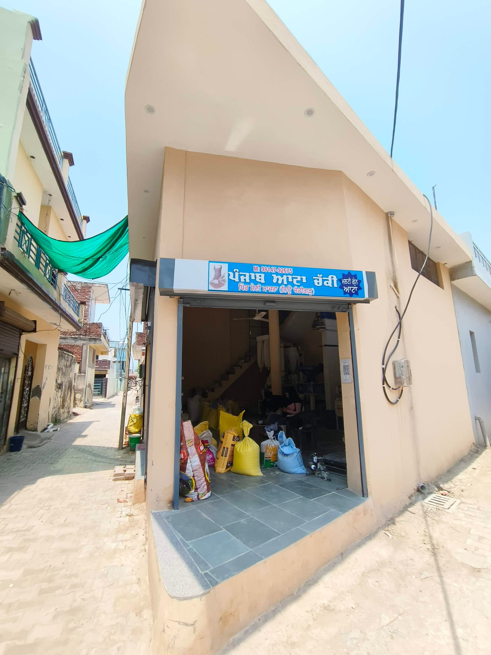 Open storefront on a narrow sunlit street, blue sign above entry and colorful bags piled outside the shop. Inside stairs are visible through the doorway.