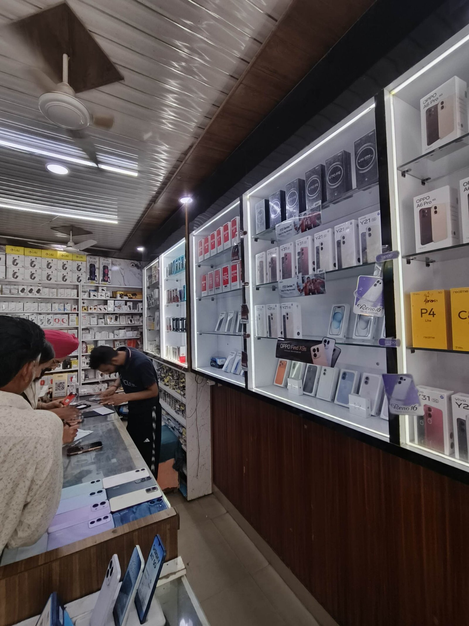 Inside a mobile phone shop with shelves of phone boxes and display phones along glass cases; customers at the counter.