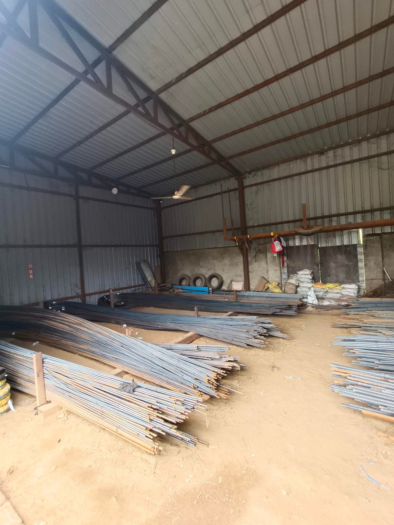 Interior of a metal workshop/warehouse with bundled steel rods stacked on the dusty floor and exposed ceiling beams at top. 0Conveys cluttered storage of construction materials.