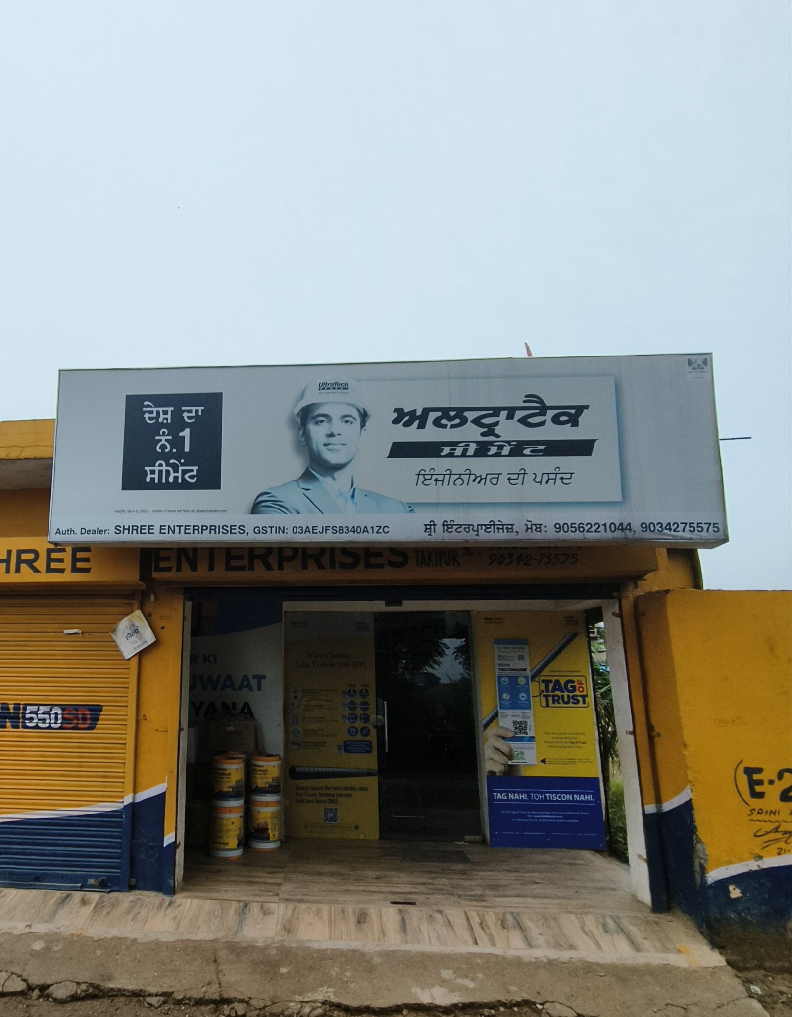 Storefront with yellow shutters and a large billboard showing a man in a hard hat, Punjab/Indian text, and entrance doors.