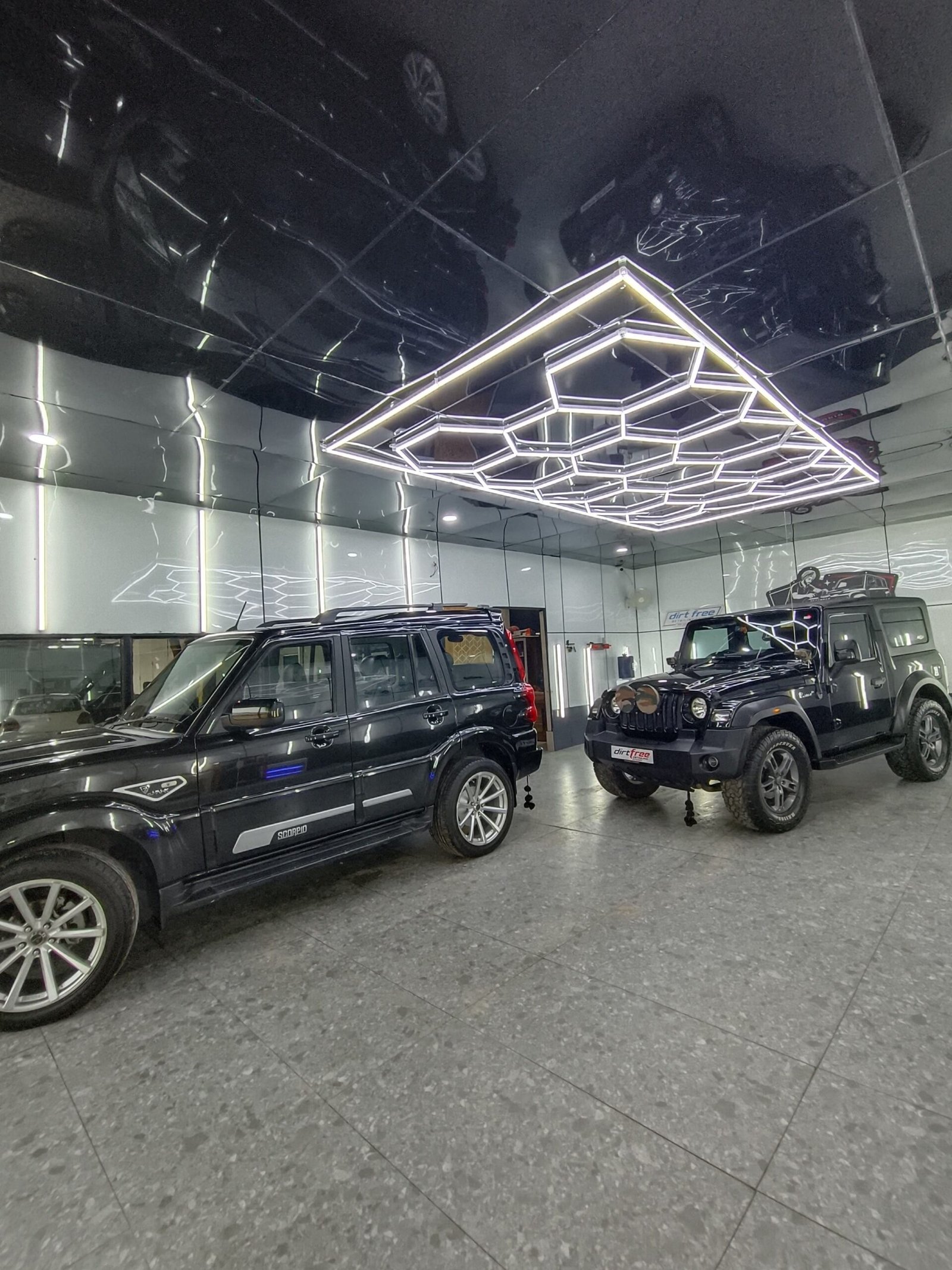 Showroom with two black SUVs (Mercedes G‑Class and Jeep Wrangler) beneath a white geometric neon ceiling fixture reflected in glossy panels on the ceiling.