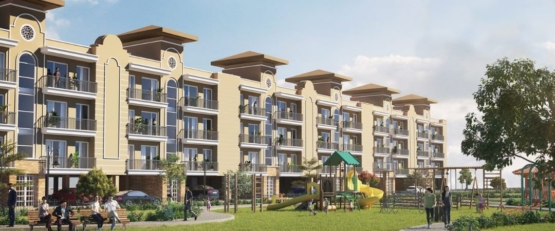 Row of beige multi-story apartment buildings with balconies around a green common area, featuring a playground and people walking and sitting nearby.