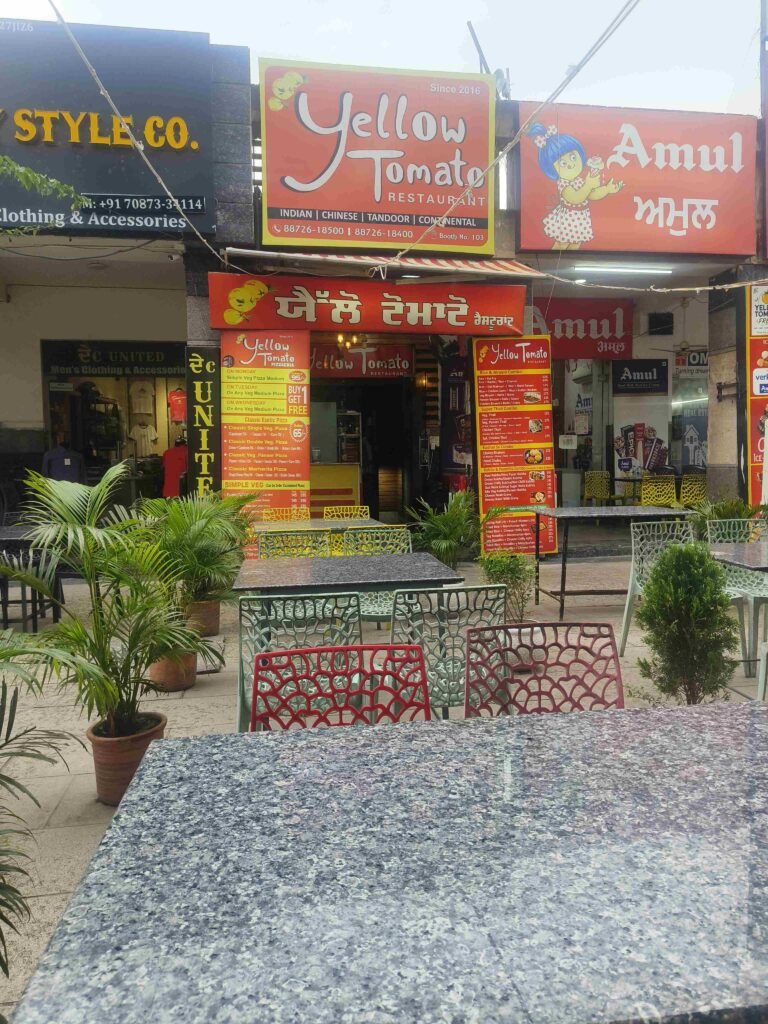 Outdoor cafe area with blue-gray granite tables and colorful red and green metal chairs, in front of Yellow Tomato restaurant storefronts and signs.