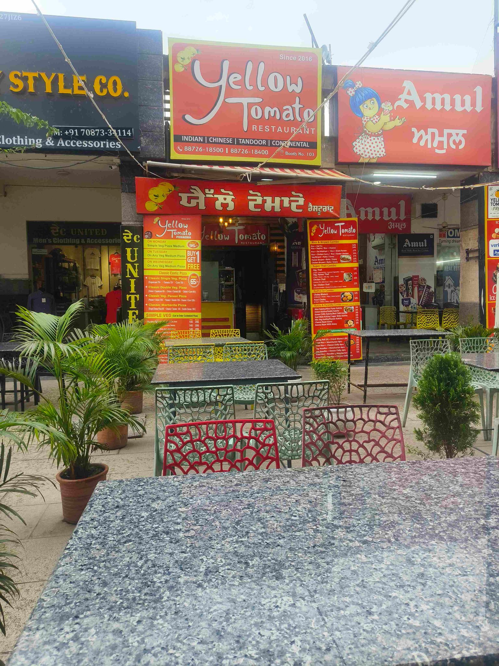 Outdoor cafe area with blue-gray granite tables and colorful red and green metal chairs, in front of Yellow Tomato restaurant storefronts and signs.