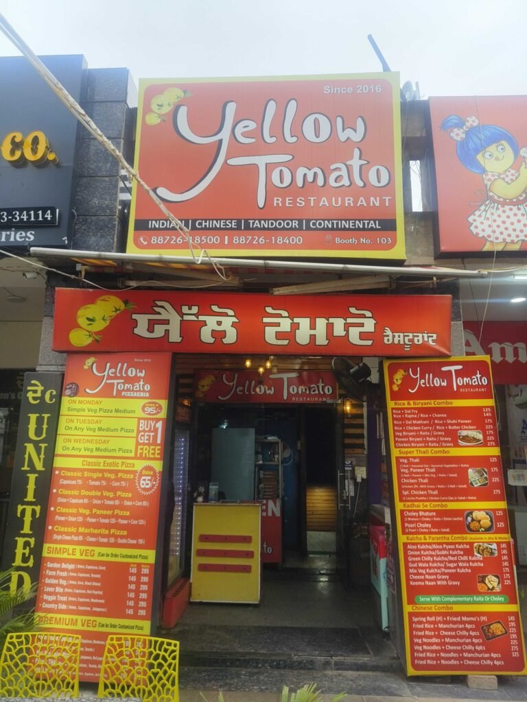 Front of Yellow Tomato Restaurant with a large orange sign reading 'Yellow Tomato Restaurant' and smaller menu boards outside, indicating Indian/Chinese cuisine.