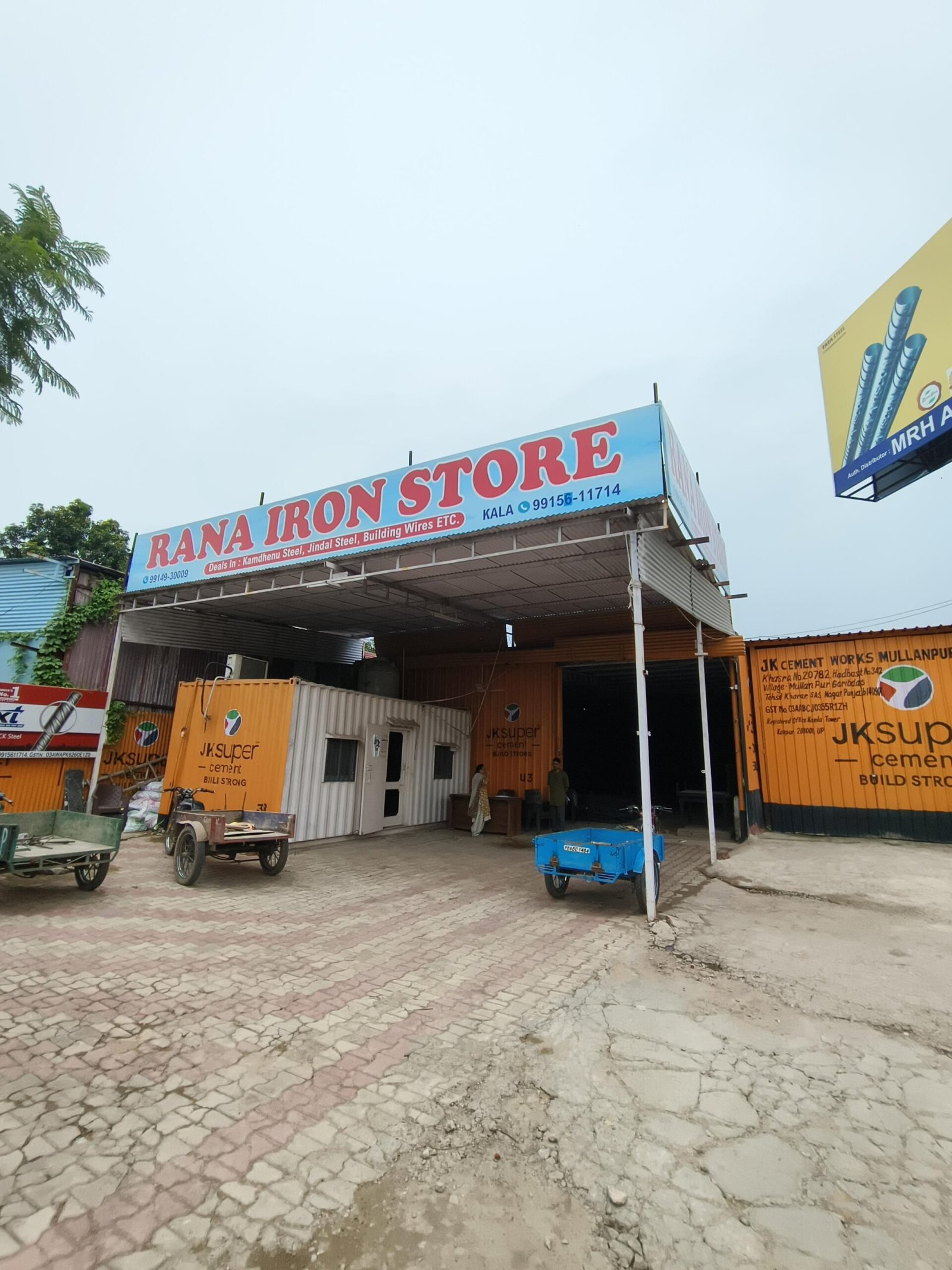 Open storefront showing Rana Iron Store sign on blue banner, with orange cement containers and carts in a construction yard nearby.