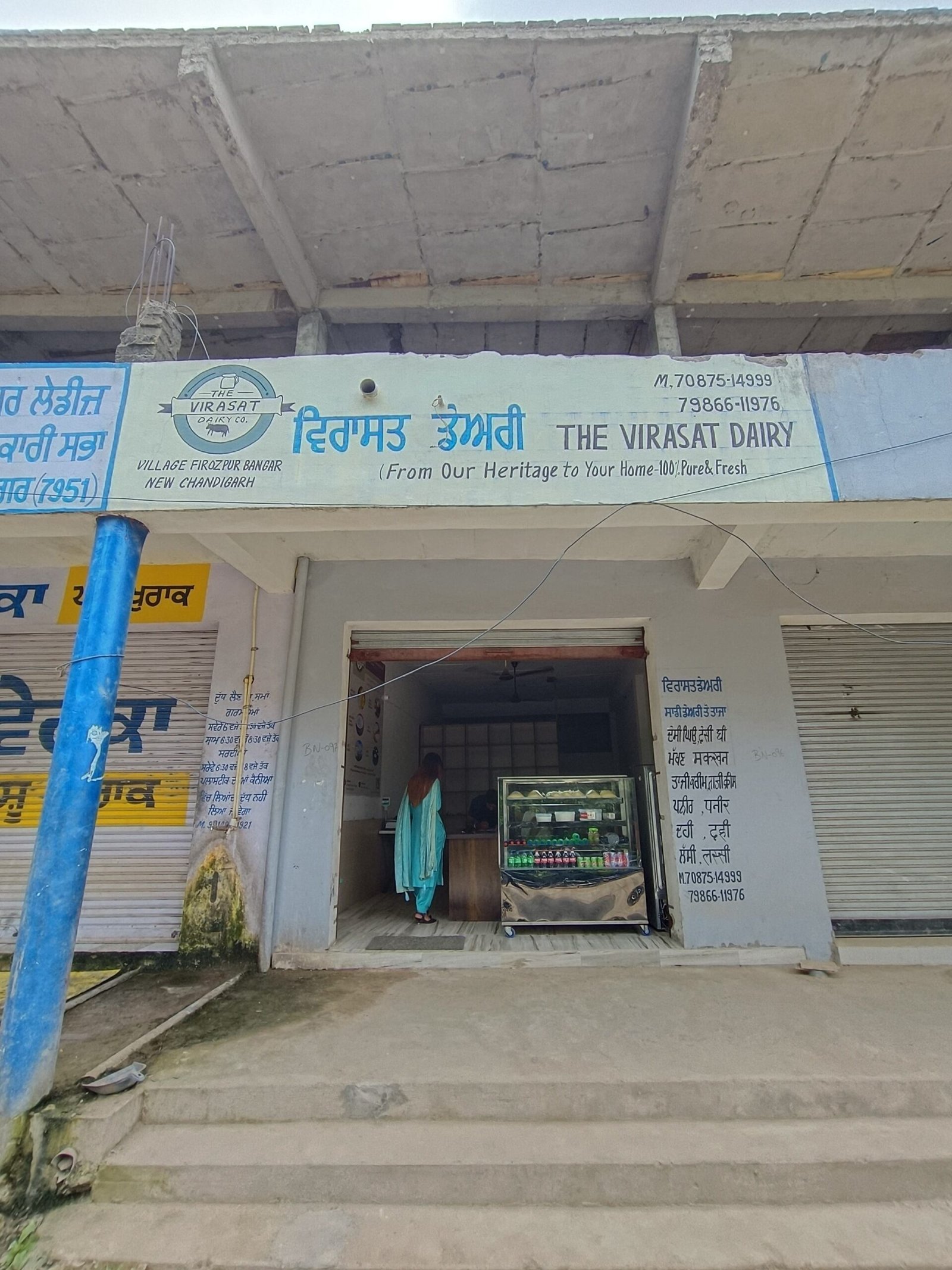 Shopfront sign reads The Virasat Dairy with a woman in a green sari at the counter inside the dairy display case.